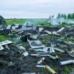 Wreckage of crashed plane scattered across field, smoke rising.