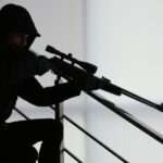 Person in dark clothing aiming a rifle indoors.