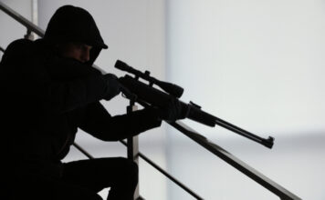 Person in dark clothing aiming a rifle indoors.