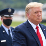 Donald Trump in suit with military personnel behind him.