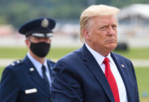 Donald Trump in suit with military personnel behind him.