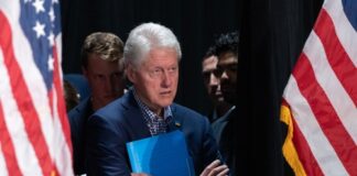 Bill Clinton standing between American flags with a folder.