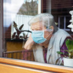 Elderly woman wearing mask, looking out window beside orchid.