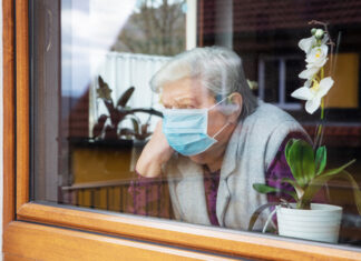 Elderly woman wearing mask, looking out window beside orchid.