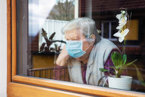 Elderly woman wearing mask, looking out window beside orchid.