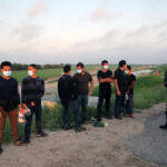 People wearing masks stand near border patrol agents outdoors.