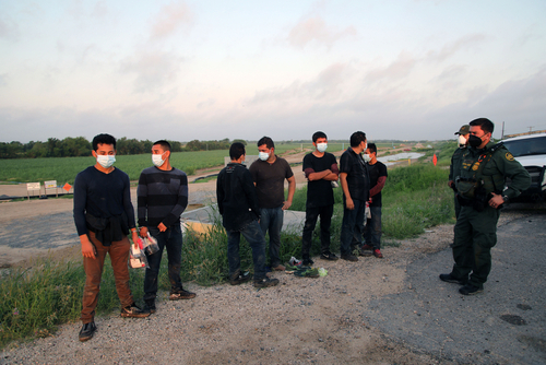 People wearing masks stand near border patrol agents outdoors.