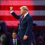 donald trump in suit raising fist at event with flag backdrop