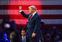 donald trump in suit raising fist at event with flag backdrop