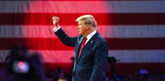 donald trump in suit raising fist at event with flag backdrop