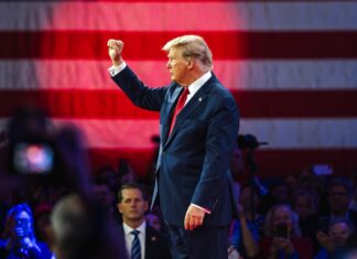 donald trump in suit raising fist at event with flag backdrop