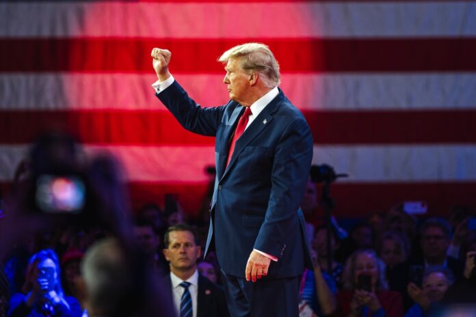 donald trump in suit raising fist at event with flag backdrop