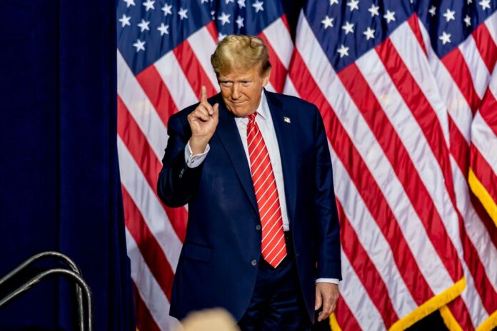 Man standing before American flags wearing suit and tie