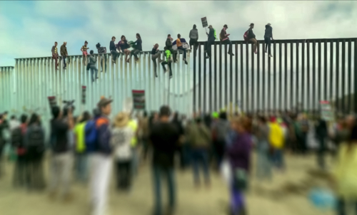 Crowd and people climbing large border wall during protest.