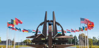 NATO emblem with flags of member countries in background.