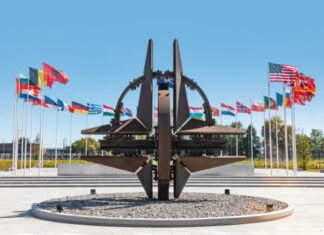 NATO emblem with flags of member countries in background.