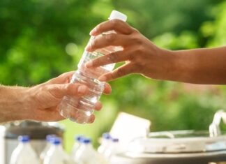 Hands passing a water bottle outdoors, lush green background.