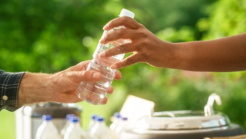 Hands passing a water bottle outdoors, lush green background.