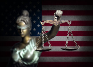 Blindfolded Lady Justice statue with scales, American flag backdrop.