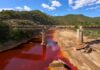 Weathered bridge over a red river with green hills.