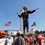 Bush-Era Swamp Creature FUNDS Cornyn’s Survival… Giant cowboy statue surrounded by crowd, American and Texas flags.