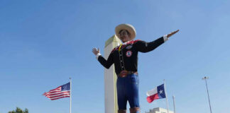 Giant cowboy statue surrounded by crowd, American and Texas flags.