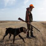A man in hunting gear walking with a dog in a field during sunset