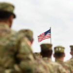 Soldiers in uniform standing, American flag in background.