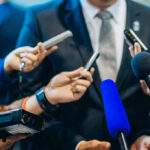 Media Flap Explodes—Death Penalty Marches On Journalists holding microphones interview a person in a suit.