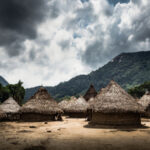 A rural village with thatched huts set against a mountainous backdrop under a cloudy sky
