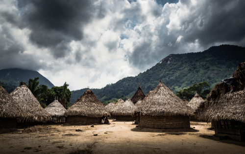 Image,From,An,Indigenous,Village,In,Guajira,,Colombia A rural village with thatched huts set against a mountainous backdrop under a cloudy sky