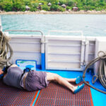 A man lying on the deck of a boat surrounded by fishing ropes with a view of the ocean