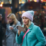 A woman in a blue coat and pink beanie speaking into a smartphone in an outdoor setting