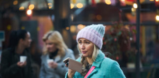 A woman in a blue coat and pink beanie speaking into a smartphone in an outdoor setting