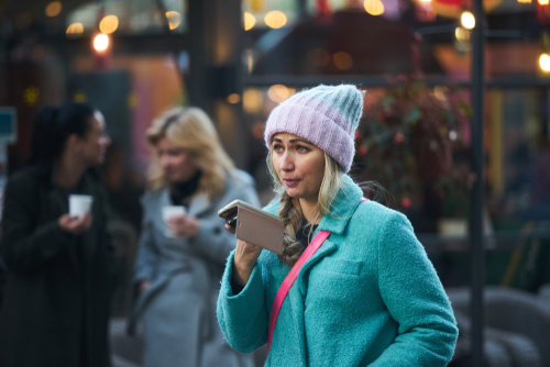 Riga,,Latvia,-,17.11.2018,Woman,In,Hat,With,Phone,In A woman in a blue coat and pink beanie speaking into a smartphone in an outdoor setting