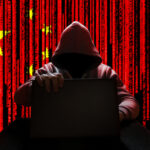 A hooded figure sitting at a desk with a laptop, against a red background featuring the Chinese flag