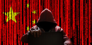 A hooded figure sitting at a desk with a laptop, against a red background featuring the Chinese flag