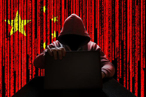 A hooded figure sitting at a desk with a laptop, against a red background featuring the Chinese flag