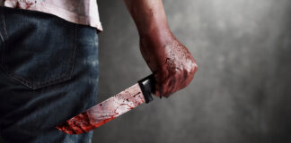 A man holding a bloody knife behind his back in a dark setting
