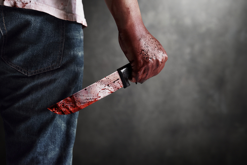 A man holding a bloody knife behind his back in a dark setting