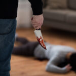 A person holding a bloody knife stands over a victim lying on the floor