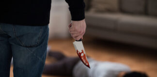 A person holding a bloody knife stands over a victim lying on the floor