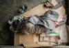 Person sleeping on cardboard with a blanket outdoors.