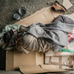 Person sleeping on cardboard with a blanket outdoors.