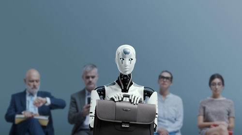 shutterstock_2268772859 Robot holding a briefcase sits among human job candidates