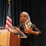 Brutal Line Boomerangs On Dems Ilhan Omar speaking at a podium with a microphone