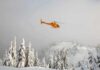 Yellow helicopter flying over snowy forested mountains