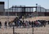 Crowd of people lining up near a border fence with barbed wire