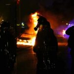 Curfew Imposed After Explosive Onslaught Riot police stand near burning cars at night