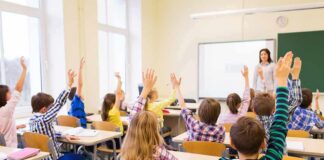 Students raise hands in a classroom while a teacher stands at the front
